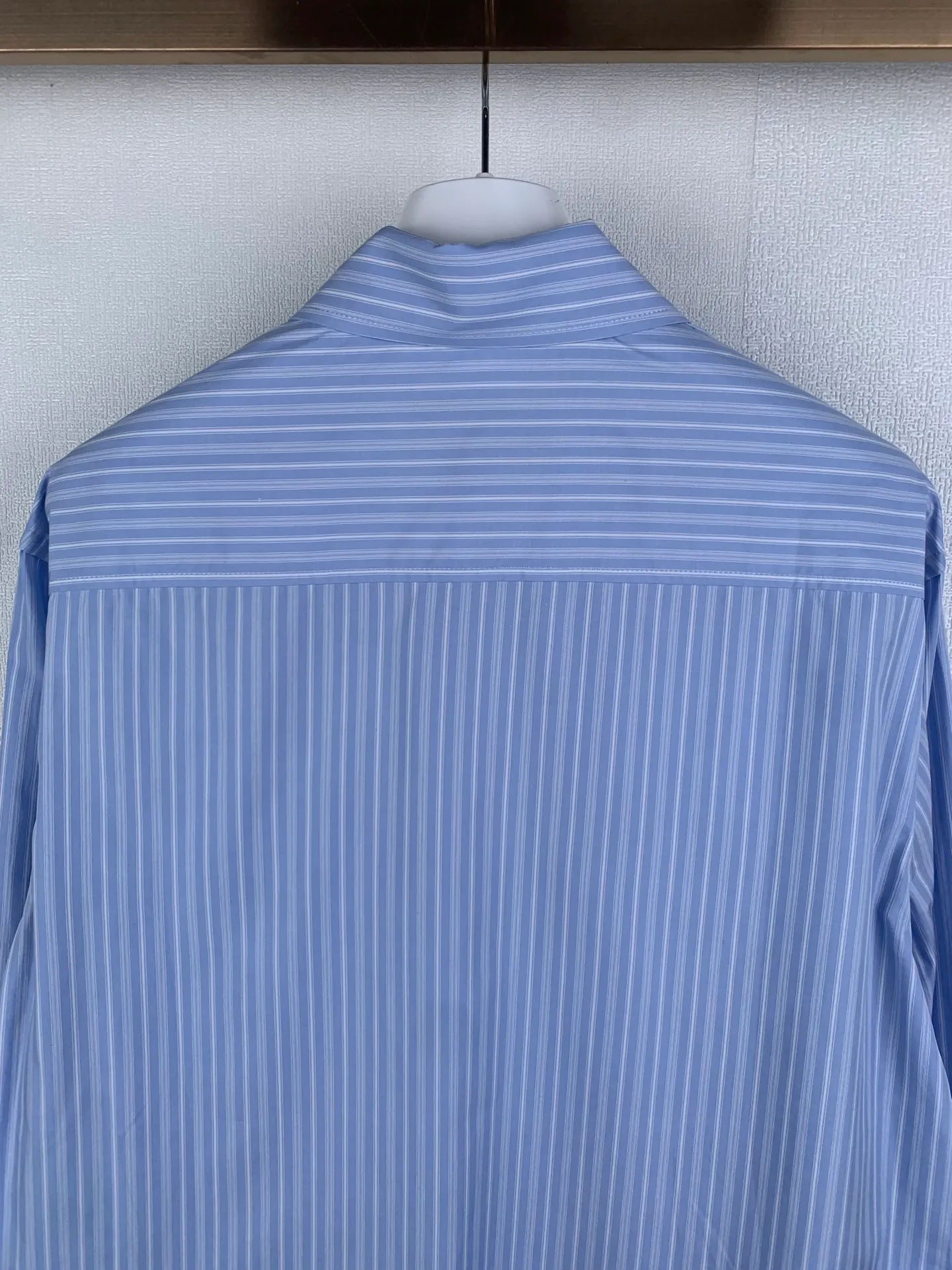 AMI embroidered love heart decorated blue and white striped long-sleeved shirt. Fabric selection: Made of 100% cotton poplin fabric. The pure cotton fiber can quickly absorb moisture and sweat, making it refreshing and not stuffy when worn for a long time. The unique plain weaving process of poplin makes the fabric both soft and skeletal, not easy to wrinkle and deform, and the upper body is neat and clean. Coupled with the fine blue and white stripes all over the body, the light color scheme is gentle and elegant, which can meet the needs of different scenes. Process 1: Accurately mark the embroidery position on the chest patch pocket to ensure that the pattern does not shift and does not destroy the overall beauty of the striped fabric. High-count embroidery threads of the same color as the fabric are selected, and flat stitch embroidery technology is used. The stitches are evenly tracked, the edges are clean and neat, and the love outline is simple and smooth. It retains recognition without irritating the skin. It is blended into the striped texture in a low-key manner to avoid an abrupt feeling, while ensuring that the embroidery threads are Wear-Resistant. 2: After the embroidery is completed, perform high-temperature ironing and thread trimming to ensure that the pattern is smooth and compliant, and it is not easy to lose thread or warp after being worn for a long time. Details: 1. The collar seat adopts double-layer reinforcement technology, which is not easy to collapse and deform after long-term wear. The collar corner has a soft arc, which can modify the neck line without being too sharp, making it neat and decent. 2. The inner side of the placket has hidden hemming treatment to avoid burrs on the fabric, while enhancing structural stability and preventing it from deforming after multiple washes. Accessories: One to one customized buttons Size: S M L XL